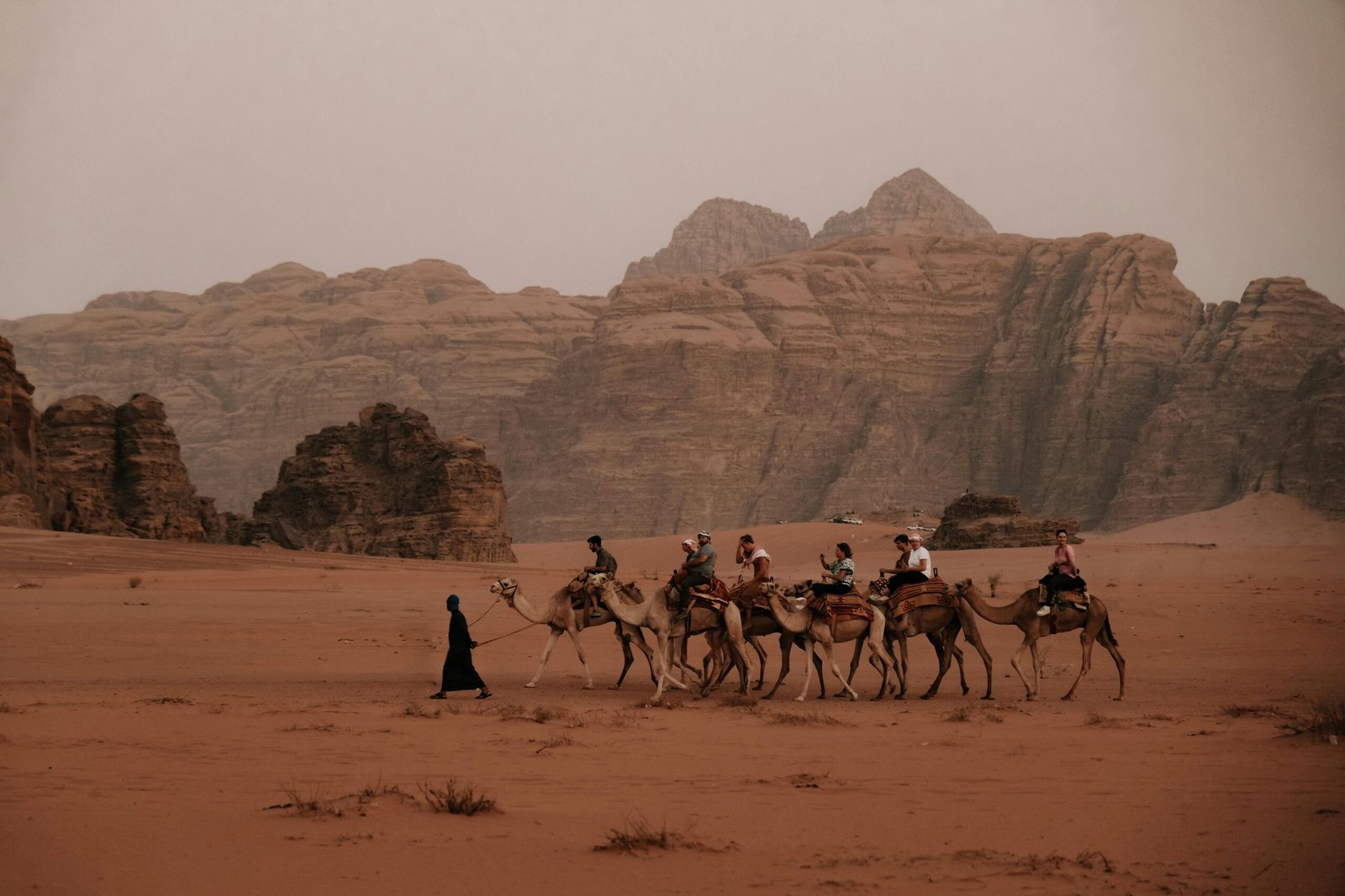 A group of tourists riding camels led by a guide in Wadi Rum, Jordan, with stunning sandstone mountains.