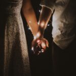 A warm and intimate close-up of a couple holding hands adorned with glowing string lights.